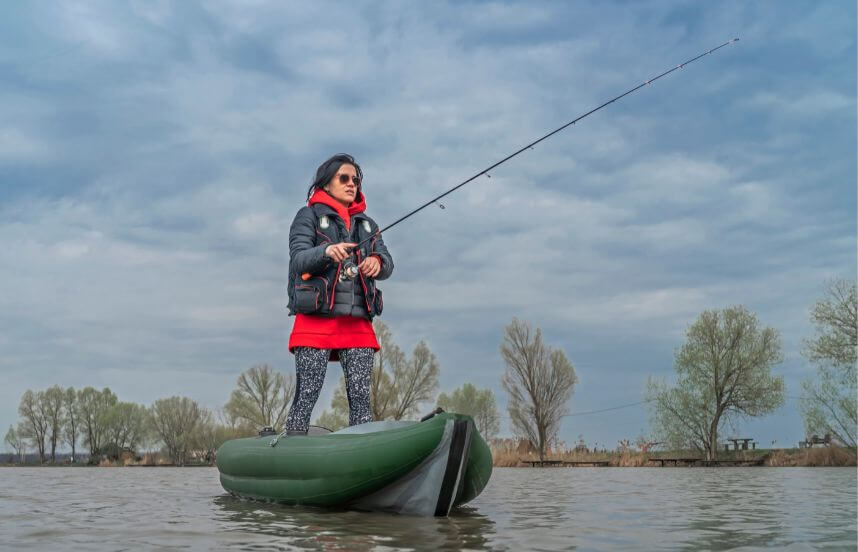 A women standing on a green inflatable kayak holding a fishing rod.